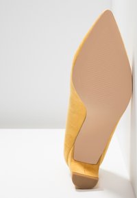 Chaussure à talon jaune en daim à bout pointu, vue par la semelle avec une surface antidérapante texturée, positionnée debout contre une surface blanche.