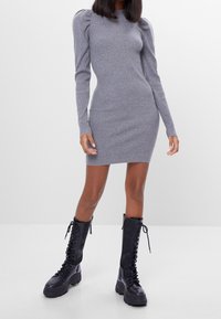 Kvinna i en figurnära grå ribbad klänning med puffärmar och knähöga svarta kängor med snörning, stående mot en neutral bakgrund.