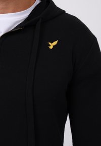 Schwarze Zip-Hoodie mit weicher Textur. Mit einem kleinen, goldenen, gestickten Vogel-Logo auf der Brust und einer Kapuze mit Kordelzug.