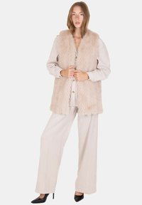 Chaleco de piel sintética en beige claro con una superficie texturizada, combinado con una blusa color crema y pantalones anchos de rayas. Zapatos de tacón altos negros.