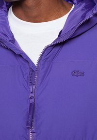 Fialová prošívaná bunda se zapínáním na zip vpředu, vybavená texturovanou látkou, nastavitelnou kapucí a výšivkou loga na hrudi.