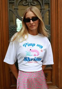 Blonde woman wearing black sunglasses, white T-shirt with "Pimp my Shrimp" text and pink shrimp graphic, and pink skirt with green spots.