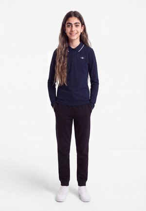 Fată cu păr lung, purtând un tricou polo albastru închis cu mâneci lungi, pantaloni negri și adidași albi, stând cu mâinile în buzunare și zâmbind.