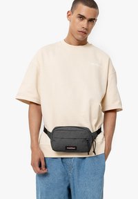 Marsupio di tessuto grigio con tasca anteriore zip, fascia nera regolabile, indossato sopra un maglione beige e jeans azzurri chiari.