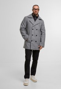 Grijze dubbelzijdige peacoat met grote zwarte knopen, voorzien van een brede kraag. Draagt over een zwarte ritssluiting jas, gecombineerd met zwarte broek en sneakers.