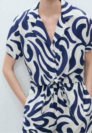 Eine Frau trägt einen kurzärmeligen, knöpfbaren Jumpsuit mit Kragen, einem gebundenen Taillengürtel und auffälligen dunkelblauen abstrakten Wirbeln auf weißem Hintergrund.