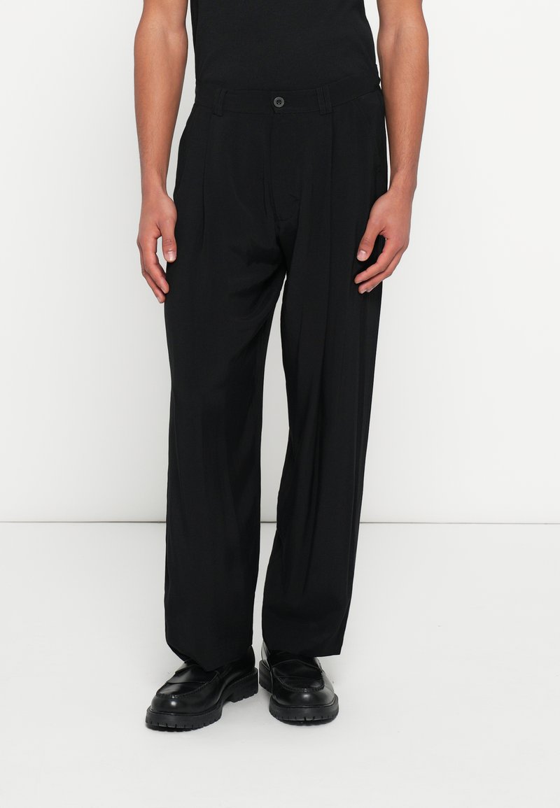 Μαύρο πλισέ παντελόνι με σχεδιασμό ίσιας γραμμής, ραμμένο σύμφωνα με το σώμα και κλείσιμο με κουμπί. Κατασκευασμένο από ελαφρύ υλικό, συνδυάζεται με μαύρα παπούτσια.