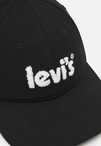 Melna beisbola cepure, izgatavota no kokvilnas auduma, ar izteiktu balto izšūto "levi's" logotipu priekšā. Noapaļota mala un tekstūrēts audums.