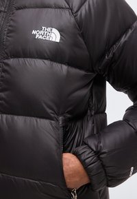 Gros plan d’une doudoune noire avec le logo The North Face sur la poitrine et une main dans la fermeture éclair de la poche latérale.