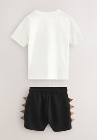 T-shirt blanc à manches courtes associé à un short noir à taille élastique avec des piques de dinosaure marron sur les côtés.