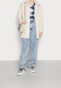 Beżowy płaszcz podszyty polarem, niebieski krótki top w romby, jasne szerokie dżinsy oraz białe trampki z czarnymi akcentami. Beżowa torba z detalem w formie łańcuszka.