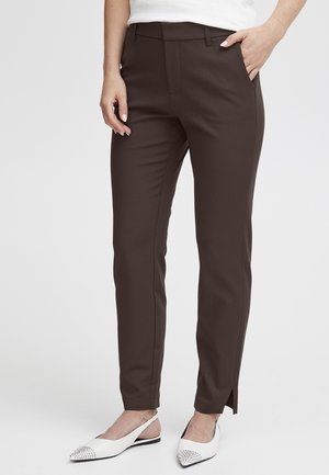 Vrouw draagt donkerbruine slim-fit broek met voorvouw en witte puntige platte schoenen met zilveren studs, staand met één hand in de zak.