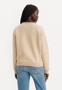 Sudadera beige con puños y cintura acanalados, que presenta un diseño clásico de cuello redondo. Combinada con jeans de mezclilla azul claro.