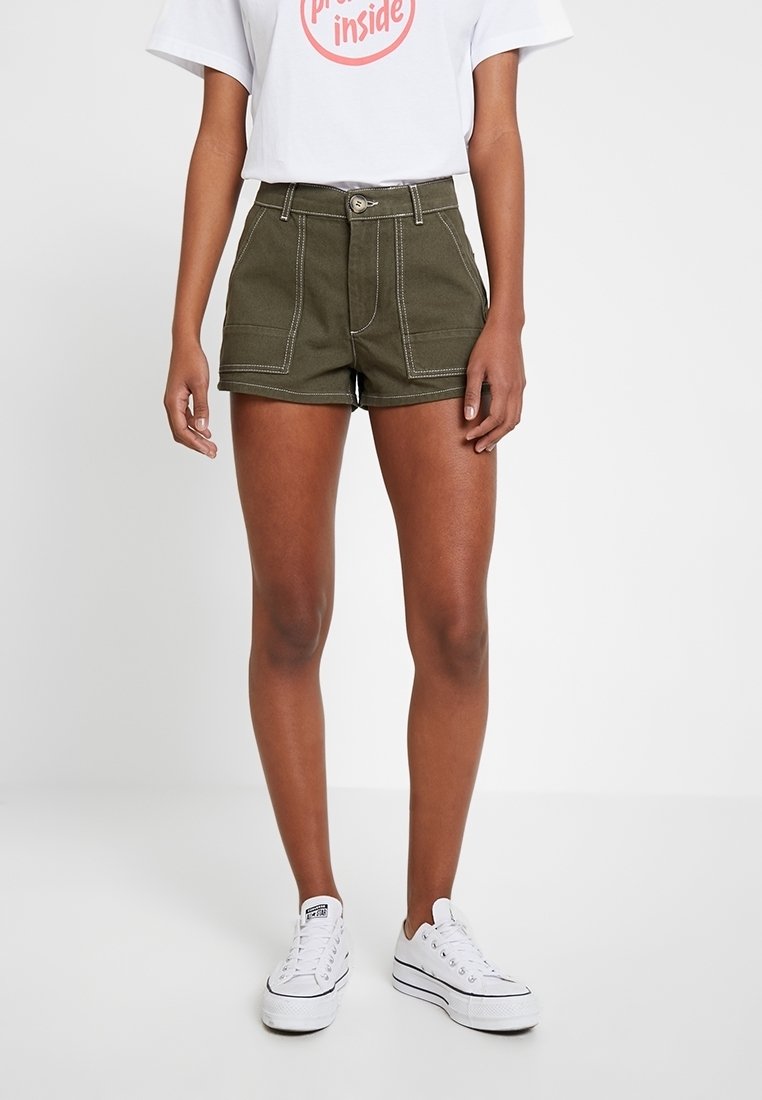 Femme portant un short taille haute vert olive avec poches avant, un t-shirt blanc à motifs graphiques, et des baskets à plateforme blanches, sur fond blanc.