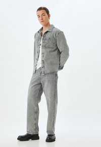 Chaqueta de denim gris y pantalones a juego con una camisa blanca debajo. Botas negras gruesas completan el conjunto. Textura suave, ajuste relajado.