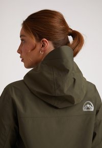 Giubbotto con cappuccio verde oliva con una texture liscia, caratterizzato da un logo montano argento sulla schiena e elementi di design minimalisti.
