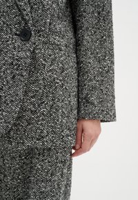 Gros plan sur une main à côté d'un blazer à motif chevron noir et blanc avec un bouton noir et un pantalon assorti.