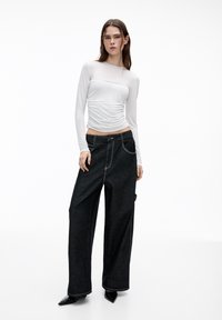 Top bianco a maniche lunghe con dettagli arricciati abbinato a jeans neri a gamba larga con cuciture rosse e tasche frontali. Tacchi neri a punta.