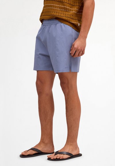Man wearing light blue shorts, brown knit top, and black flip-flops, standing against a white background, hands relaxed by sides.