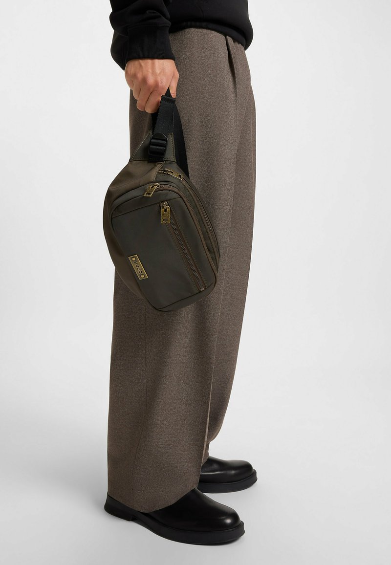Borsa in tessuto verde oliva con hardware dorato, chiusura con zip superiore e tracolla regolabile, tenuta in una mano accanto a pantaloni beige su misura e scarpe nere.