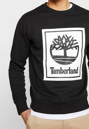 Mężczyzna w czarnej bluzie z białym logo Timberland z stylizowanym drzewem i napisem na piersi, widoczne beżowe spodnie.