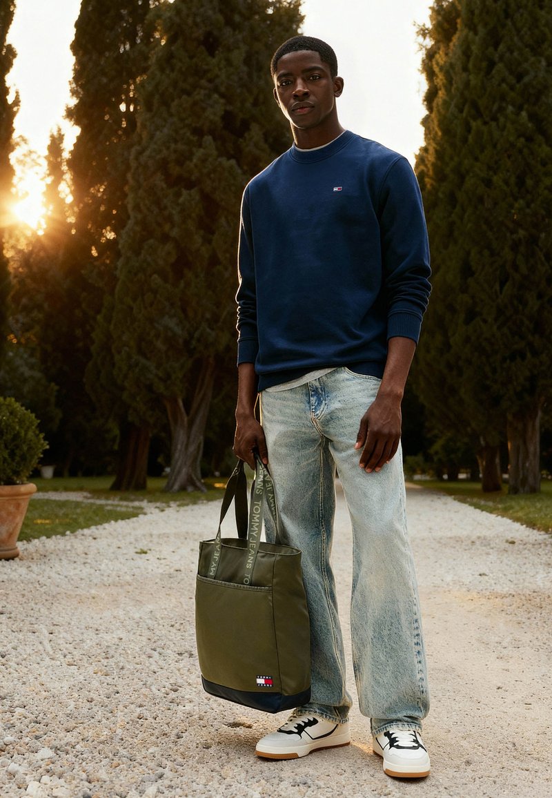 Ung mand står på grussti mellem høje træer ved solnedgang, iført marineblå sweatshirt, lyse jeans, hvide sneakers og holder en grøn mulepose.