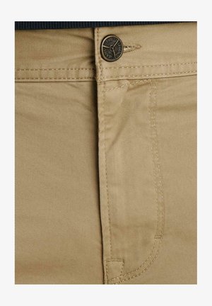 Tanové nohavice v detailnom zábere, na ktorých je kovové gombík s gravírovaným nápisom "PME Legend Jeans" a viditeľné švy na prednom vrecku.