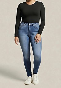 Kobieta w obcisłej czarnej bluzce z długim rękawem, niebieskich obcisłych dżinsach z wysokim stanem i białych tenisówkach, stojąca na jednolitym tle.