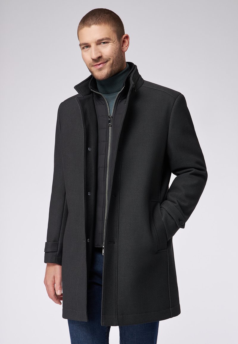 Schwarzer Wollmischmantel mit strukturiertem Muster, Knopfleiste und seitlichen Taschen. Darunter getragen mit einer schwarzen, mit Reißverschluss versehenen Innenjacke.