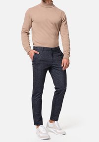 Beige Rollkragenpullover und dunkelblaue gemusterte Hose, kombiniert mit hellen Sportschuhen. Figurbetonter Schnitt mit schmal zulaufenden Beinen.