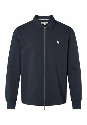 Marine blauwe ritsjack in zachte stof, met een kraag, lange mouwen en een klein wit logo op de borst. De rits loopt verticaal over de voorkant.