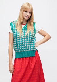 Turkusowa i szara dzianinowa kamizelka z dekoltem w serek, z fakturowanym wzorem i prążkowanym dołem, noszona na białym T-shircie. Połącz z czerwoną spódnicą.