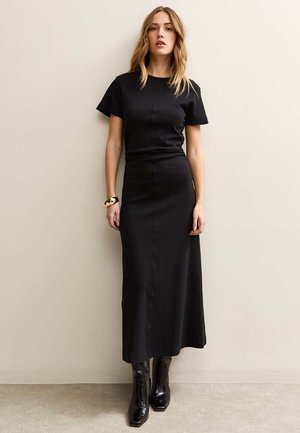 Donna in piedi contro una parete beige che indossa un abito lungo nero aderente a maniche corte, stivaletti neri e un braccialetto d'oro al polso sinistro.