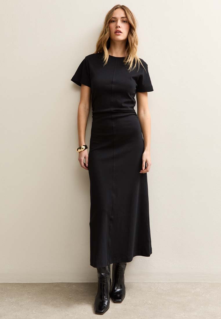 Donna in piedi contro una parete beige che indossa un abito lungo nero aderente a maniche corte, stivaletti neri e un braccialetto d'oro al polso sinistro.