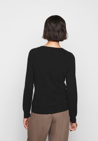 Maglione nero a maniche lunghe con scollo rotondo, realizzato in materiale morbido. Presenta un design aderente e un orlo a coste. Indossato sopra pantaloni marroni.