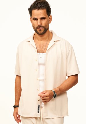 Mann mit Bart, der ein offenes beiges Kurzarmhemd über einem weißen Tanktop, einer silbernen Uhr und Halsketten vor einem schlichten Hintergrund trägt.