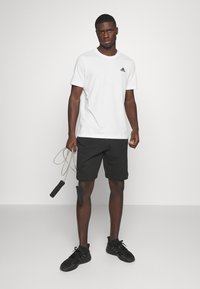 Bela kratka majica s kratkimi rokavi in črnim logotipom, v kombinaciji s črnimi športnimi hlačami in črnimi športnimi copati, drži skakalno vrv.