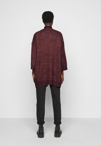 Burgundská oversized pletená kardigan s texturovaným povrchem, spadlými rameny a ribbovaným lemem, v kombinaci s černými kalhotami a robustními botami.