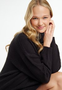 Maglione nero a maglia con un motivo testurizzato, collo alto e polsini a costine. La modella è seduta, mentre tocca delicatamente il suo viso. Sfondo uniforme.