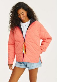 Korálová prošívaná bunda s černým límcem, zip zvýrazněný barevnými třásněmi a bočními rozparky. V kombinaci s džínovými šortkami.