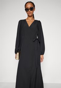 Rochie neagră, cu mâneci lungi și puffate, și o curea la talie. Textură fină, decolteu în V, și se termină la nivelul gleznelor. Stilizată cu ochelari de soare.