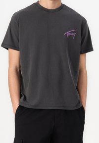 Mörkgrå bomullströja med rund halsringning. Har en lila och rosa logotyp med texten "Tommy" på vänster bröst. Korta ärmar.