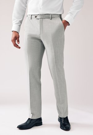 Homme portant un pantalon de costume gris clair avec des chaussures noires et une chemise blanche à manches longues, debout, une main dans la poche.