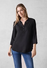 Sort, langærmet bluse med V-hals og knaplukning, lavet af glat stof. Kombineret med blå tætsiddende jeans.