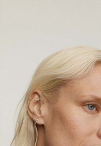 Primer plano de una persona con cabello rubio platino y ojos azules, que lleva un aro de oro delgado adornado con pequeñas piedras preciosas.