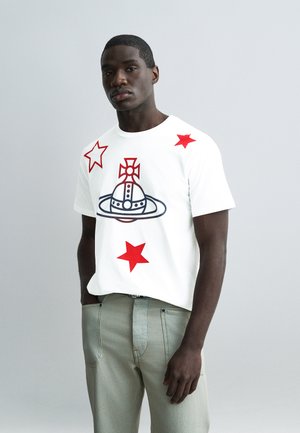 Junger Mann mit dunkler Haut, der ein weißes T-Shirt mit roten Sternen und einem schwarz-roten Globus-Emblem trägt, kombiniert mit hellgrauen Hosen.