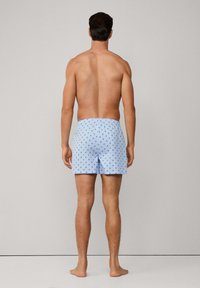 Svijetloplavi bokserice s malim crnim uzorkom križa, elastična stražnja traka i opušteni kroj. Nose ih muškarac bez majice, prikazan iz pozadine.