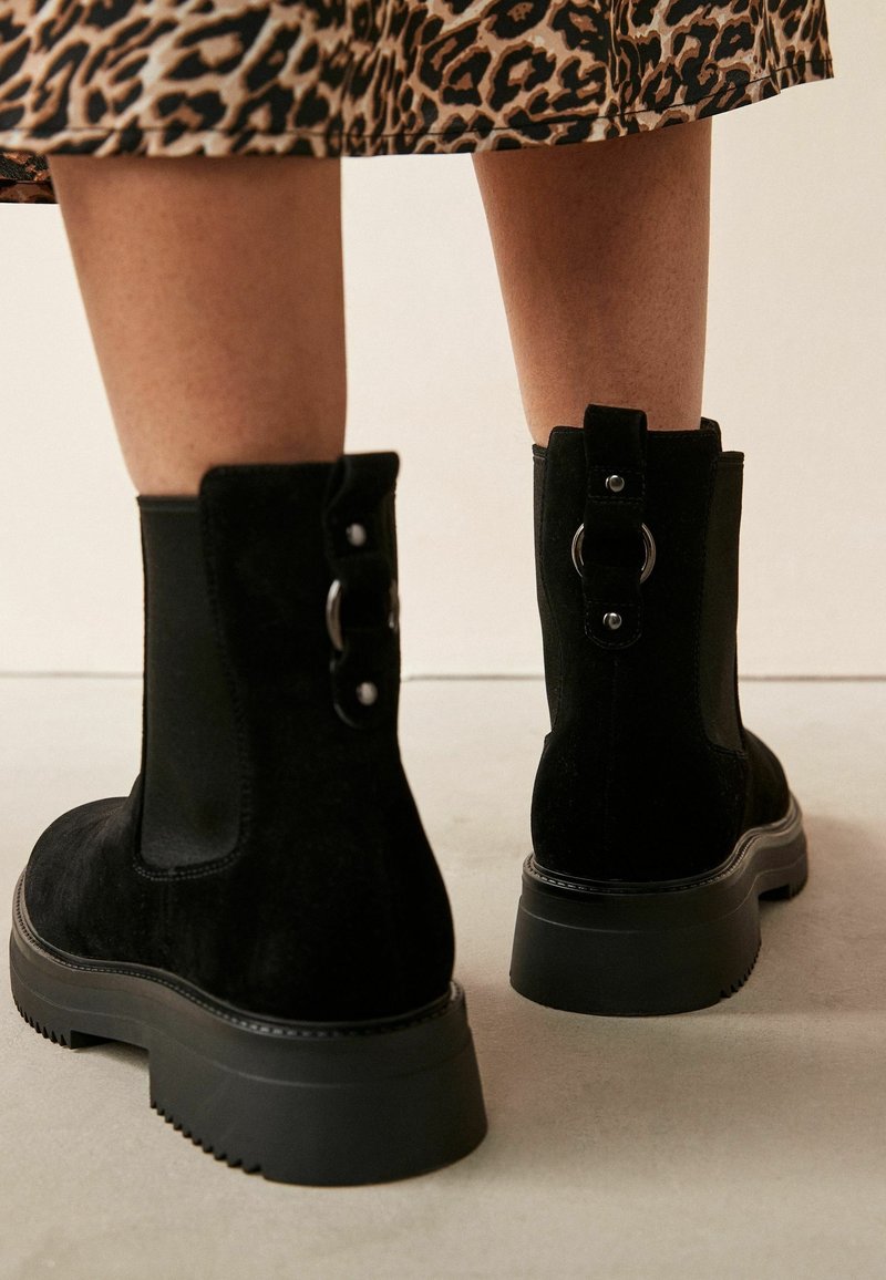 Bottes cheville en daim noir avec semelles épaisses et détails anneaux en métal, portées avec une jupe imprimée léopard, vue de dos.