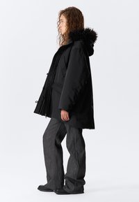 Parka neagră cu glugă căptușită cu blană, având un fermoar frontal și buzunare. Asociată cu pantaloni largi gri texturat și încălțăminte neagră.