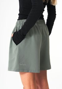 Grüne Shorts mit glatter Textur, elastischem Bund, Seitentaschen und einem Logodetail nahe dem Saum, kombiniert mit einem schwarzen Langarmoberteil.